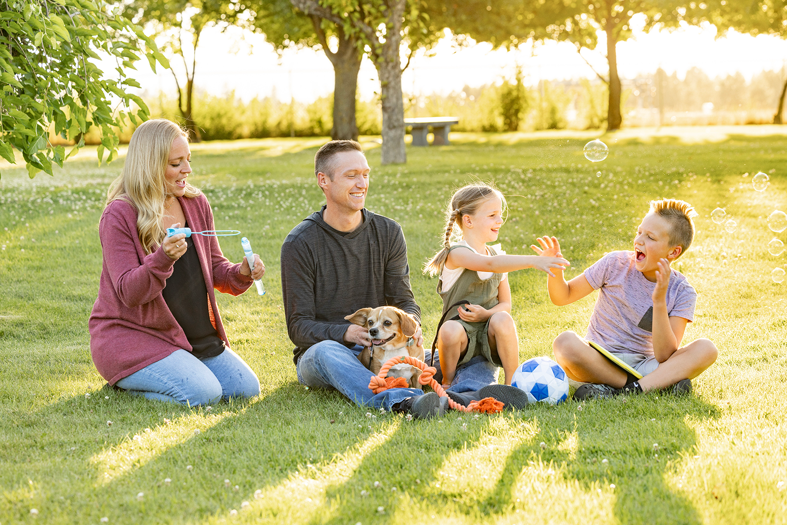 Family of four and their dog sitting inf resh grass at a playground blowing bubbles together and laughing.